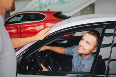 Beyaz arabanın içinde otururken yakışıklı ve memnun genç erkekler ve Yöneticisi bakın. O gülümsüyor. Adam el ele tutuşsun direksiyon simidi üzerinde. Beyaz araba ve müşteri bakmak yanında sakallı Yöneticisi standları.