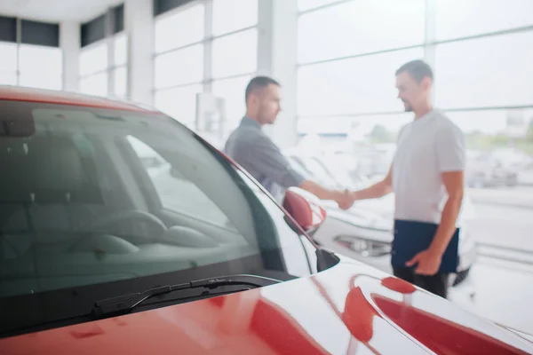 Cliente e venditore stanno dietro l'auto rossa e stringono la mano. Hanno un accordo. Il tizio con la camicia bianca tiene il tablet. Sono all'interno del negozio di auto. . — Foto Stock