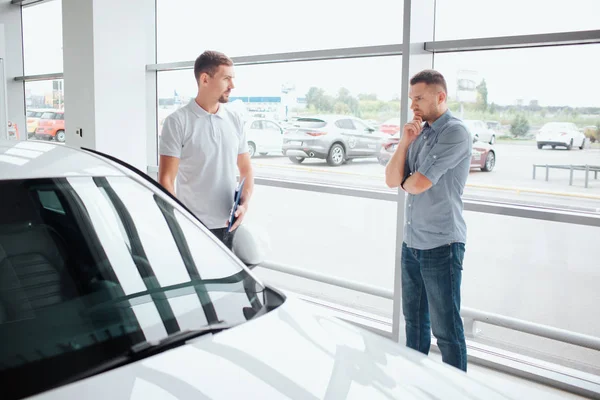 Serious and thoughtful young man stand in front of white car. He is thinking. Guy in white shirrt points on car. He looks at customer. Young man is serious.
