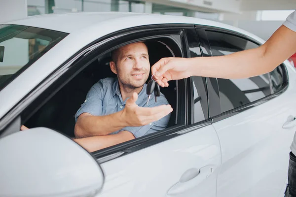 Ευτυχής κάτοχος της συνεδρίασης λευκό αυτοκίνητο στο εσωτερικό του και να κοιτάξουμε manager που δίνει το κλειδί σε αυτόν. Νεαρός άνδρας τεντώνει το χέρι του. Το άλλο είναι στο τιμόνι. — Φωτογραφία Αρχείου