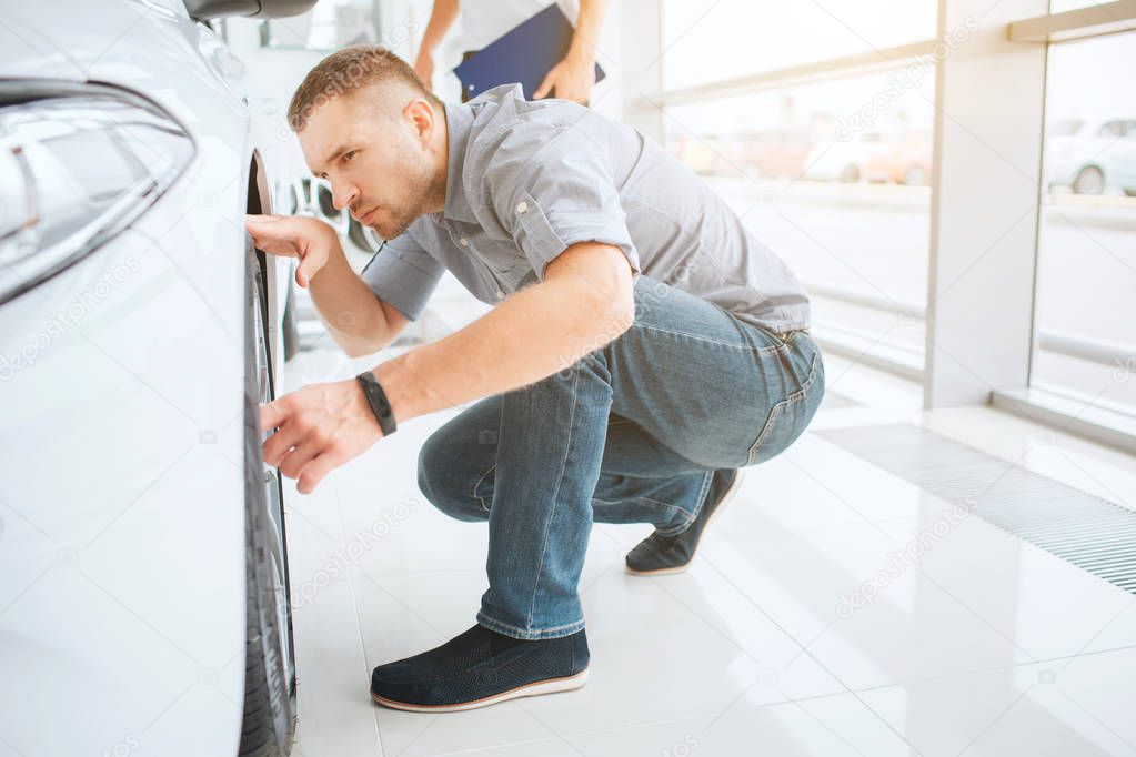 Picture of young man sitting in squad position and look at space between body of car and wheel. He is attentive to detailes. Seller stands behind him. They are inside in bright building.