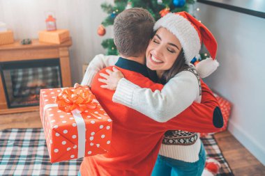 Genç kadın kucaklama adam güzel ve neşeli resmi. O gülümsüyor ve gözleri kapalı tutar. Kırmızı kutu hediye ve aşınma Noel şapka modeli tutar. Adam har kırmızı kazak.