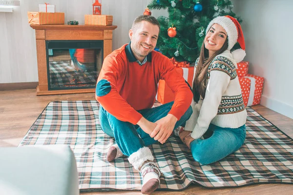 Photo d'un jeune couple heureux assis ensemble sur une couverture. Ils sourient et regardent la caméra. Les jeunes hommes portent un pull. Femme a chapeau de Noël . — Photo