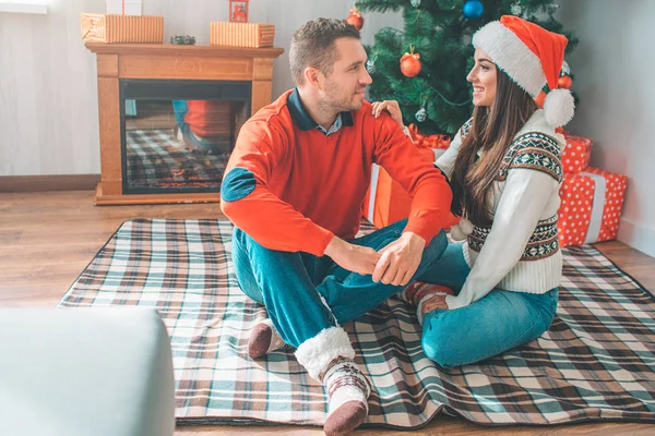 Pareja alegre sentados juntos en la manta en el suelo. Una joven toca a un tipo con la mano y míralo. Ella sonríe. Joven mírala con deificación. Mujer lleva sombrero de Navidad . — Foto de Stock