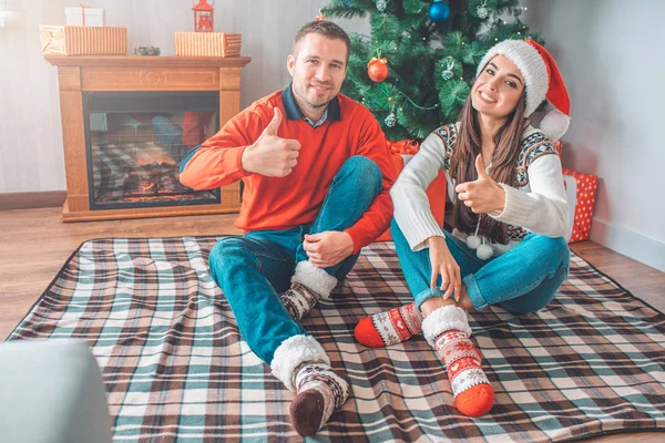 Positive picture of young couple sitting on blanket on floor and pose on camera. They keep big thumbs up and smile. People hold legs crossed.