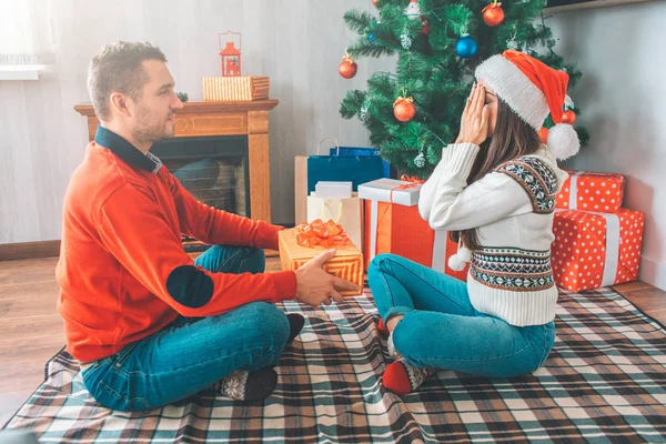 El joven se sienta frente a la mujer y la mira. Él tiene presente. La joven mantiene los ojos cerrados con las manos. Lleva suéter y sombrero de Navidad. Hay un árbol con regalos detrás de ellos . — Foto de Stock