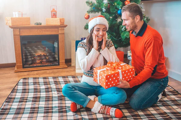 Mujer joven muy feliz y emocionada se sienta en la manta y mira al presente. Mantiene las manos en las chicas. El modelo está contento. El joven se sienta a su lado y tiene presente. La mira y sonríe. Chico feliz . — Foto de Stock