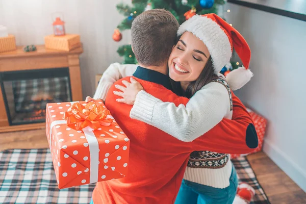 Bonito y alegre cuadro de mujer joven abrazar al hombre. Sonríe y mantiene los ojos cerrados. Modelo sostiene caja roja con regalo y usar sombrero de Navidad. Chico har suéter rojo . — Foto de Stock