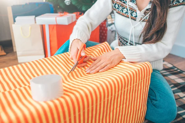 Corte la vista de la mujer sentada en el suelo y la caja de embalaje con papel naranja. Ella corta cinta con tijeras. Chica lo hace muy preciso . — Foto de Stock
