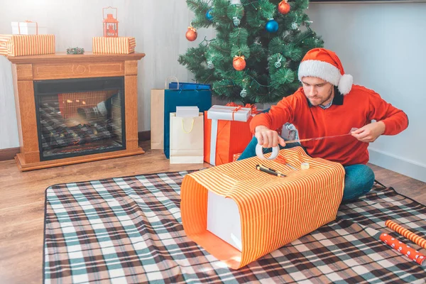 L'homme sérieux et concentré est assis sur le sol et utilise du ruban adhésif pour emballer la boîte cadeau. Il a l'air confiant. Il y a un couteau sur la boîte . — Photo