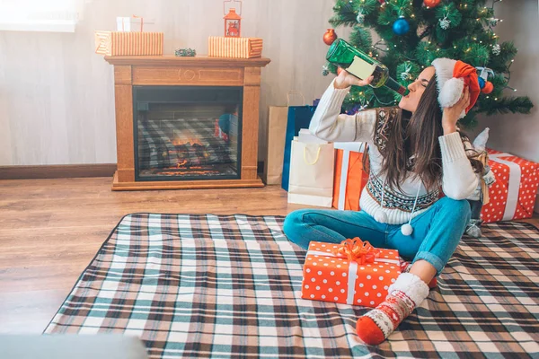 Une jeune femme fait couler de l'alcool dans une bouteille verte. Elle s'assoit sur le sol et tient la tête à la main. Il y a une boîte rouge avec un cadeau à côté de sa jambe . — Photo