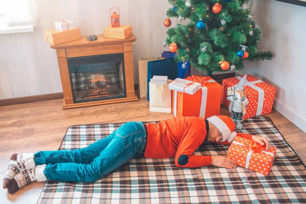 Un homme endormi couché sur le sol. Il est fatigué. Il a un pull et un chapeau de Noël. Il y a du rouge à côté de lui. . — Photo