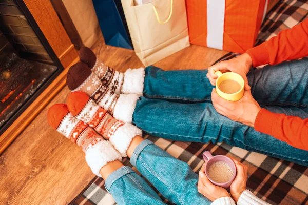 Вырежьте вид мужчины и женщины, сидящих рядом с камином и держащих чашки с горячим напитком. Они носят джинсы и рождественские носки. . — стоковое фото