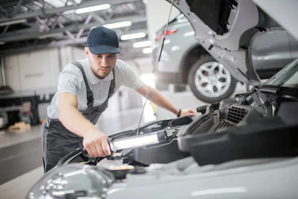 El hombre serio y concentrado se para en la carrocería abierta del coche y los detalles de la reparación en ella. Está calmado y concentrado. El tipo tiene la llave en la mano. Lo usa. . — Foto de Stock