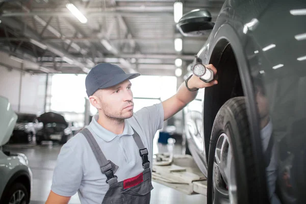 Un joven trabajador se para cerca del coche y sostiene las luces. Es serio y está concentrado. Joven mira el espacio entre el cuerpo del coche y el neumático . — Foto de Stock