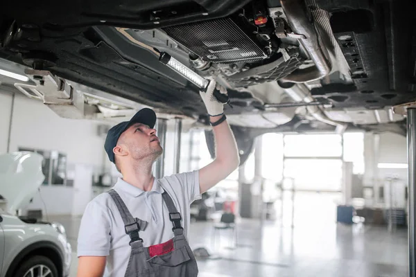 Young man in uniform stands underneath car and looks up. He holds light in hand. Guy is serious and concentrated. He works. — Stock Photo, Image