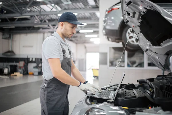 Imagen de tipo trabajador joven en el teclado. Mira la pantalla del portátil. El tipo está haciendo diagnósticos para el coche. Comprueba su estado. . — Foto de Stock