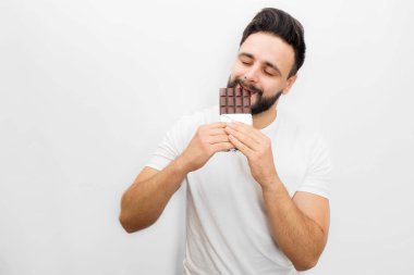 Genç sakallı adam yeme lezzetli çikolata resmi. O ısırık gözleri kapalı. Beyaz arka plan üzerinde izole.
