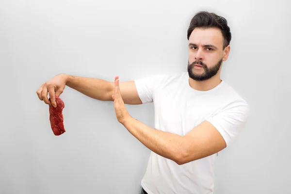 El joven preocupado se para y sostiene un pedazo de carne. Es malo. Joven muestra el símbolo de parada con la mano. Mira y posa en cámara. Aislado sobre fondo blanco . — Foto de Stock