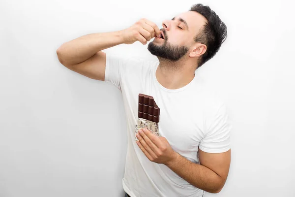 Obrázek plný radosti. Mladý vousatý muž stojany a eats čokoláda. On dává kus do úst. Chlap drží zavřené oči. Člověk je koncentrovaný. Izolované na bílém pozadí. — Stock fotografie
