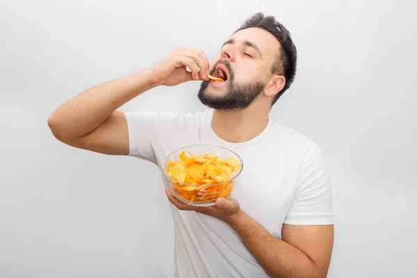 Man in een wit overhemd staat en eet chips. Hij zet het in de mond met de hand. Man heeft ook een kom met hen in een andere hand. Haar heerlijk. Geïsoleerd op witte achtergrond. — Stockfoto