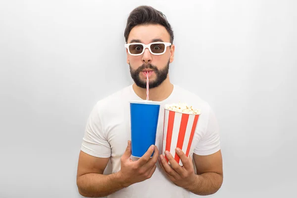 Confident young man looks on camera through movie glasses. He holds glass of coke nad popcorn in both hands. Young man drinks through straw. Isolated on white background. — Stock Photo, Image