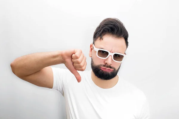 Sad and unhappy bearded young man stand and hold his big thumb down. He dislikes movie. Guy keeps cheeks blowed. Isolated on white background, — Stock Photo, Image