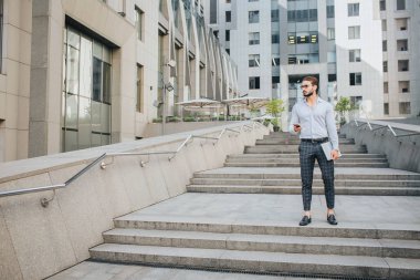 Stand ve poz ciddi ve yakışıklı genç adam. Sağa doğru görünüyor. İşadamı telefonu ve laptop elinde tutar. O binaları ile çevrilidir.