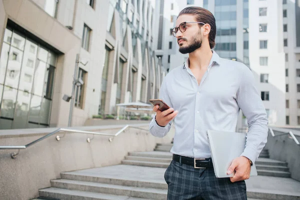 Retrato horizontal de un joven atractivo de pie en las escaleras y mira a la izquierda. Se ve moderno y elegante. El tipo tiene el portátil y el teléfono en las manos. El joven parece serio. . — Foto de Stock