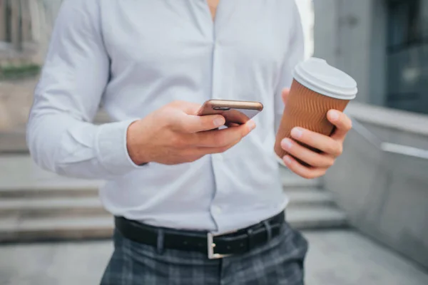 Cut view of body of young businessman stands and holds cup of coffee and phone. He is on steps. — Stock Photo, Image