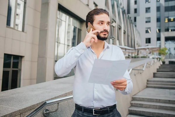 Handsome and stylish businessman stands and poses. He talks on phone and look to right. Guy holds piece of paper in left hand. Young man stands on steps. — Stock Photo, Image