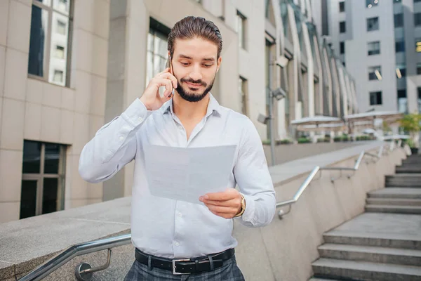 Handsome and positive young man stands on steps. He holds pece of paper and smiles. Businessman talks on phone. He looks at document. — Stock Photo, Image