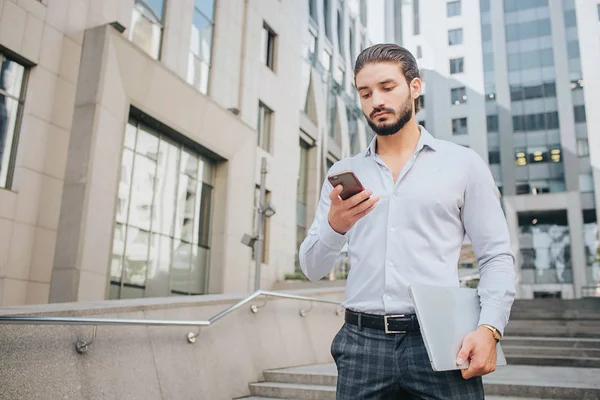 Calm and concentrated young businessman stands on steps and looks at phone. He holds laptop in left hand. Guy is surrounded with buildings. — Stock Photo, Image