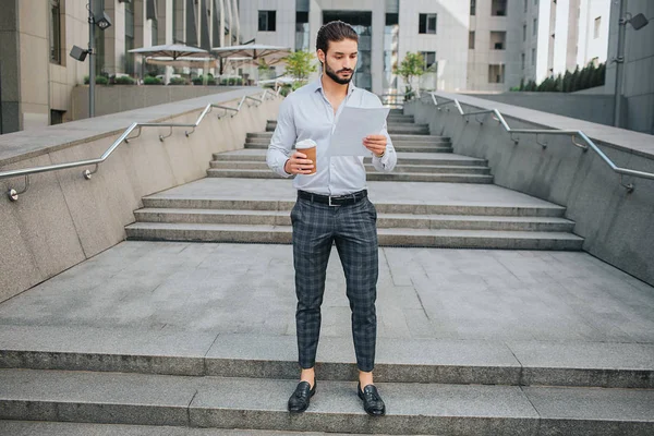 Consentrated and stylish young man stand on steps and looks at document he holds in hand. Guy is serious and calm. He has cup of coffee in right hand. Young man is surrounded with building. — Stock Photo, Image