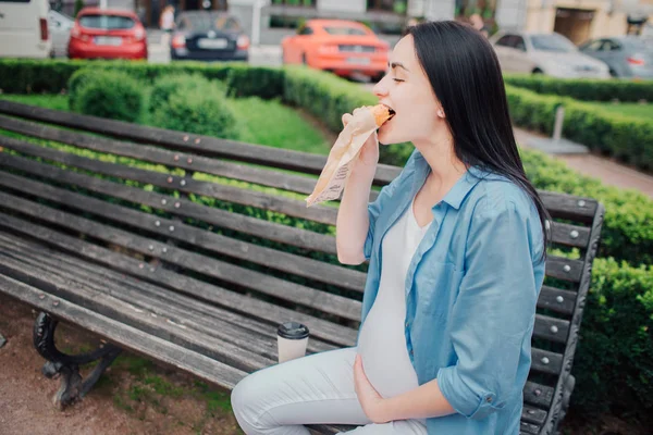 Retrato de um cabelo preto feliz e mulher grávida orgulhosa em uma cidade ao fundo. Ela está sentada em um banco da cidade e comendo uma torta ou sanduíche. Mãe expectante com fome tem uma sensação de fome — Fotografia de Stock