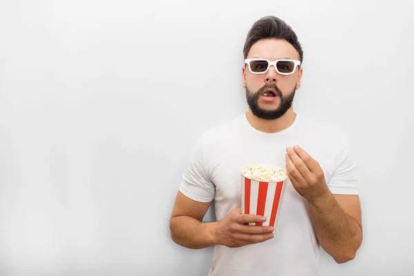 Excited and amazed young man stands and looks forward on camera. He wears movie glasses. Guy holds bucket of popcorn. His mouth is opened. Isolated on white background. — Stock Photo, Image