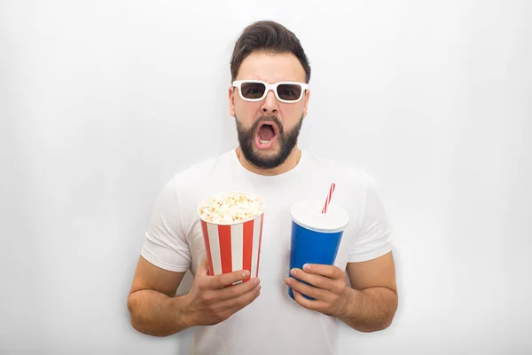 Excited man keeps his mouth opened. He holds bucket of popcorn and glass of cola at the same time. Guy wears movie glasses. Isolated on white background. — Stock Photo, Image