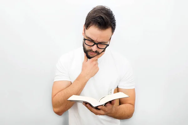 Ragazzo premuroso con gli occhiali in piedi e leggere un libro. Lo tiene nella mano sinistra. Un altro sotto il mento. Isolato su sfondo bianco . — Foto Stock