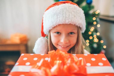 Küçük ve güzel kızın holding resmi hediye kutusu okumak ve kamera görünüyor. O mutlu. Kız Noel şapka kafasına takıyor. Ağaç arkasında.