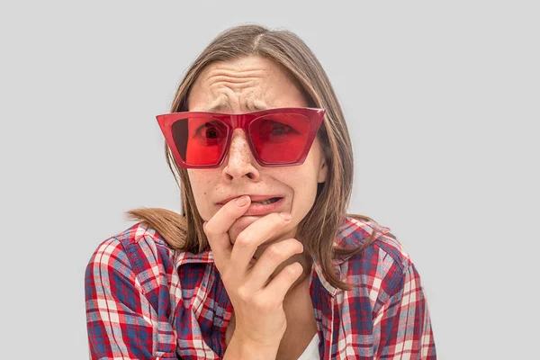 Martwię się i niezadowolony, że młoda kobieta wygląda na kamery przez czerwone okulary przeciwsłoneczne. Ona trzyma jedną rękę na usta i buty jej emocje. Na białym tle na szarym tle. — Zdjęcie stockowe