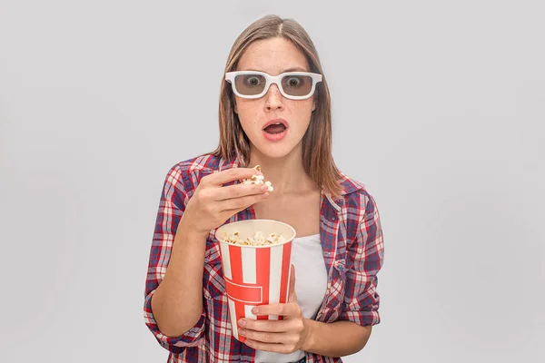 Amazed young woman keeps mouth opened and looks through glasses. She holds box of popcorn in one hand and handful of it with another one. Isolated on grey background. — Stock Photo, Image