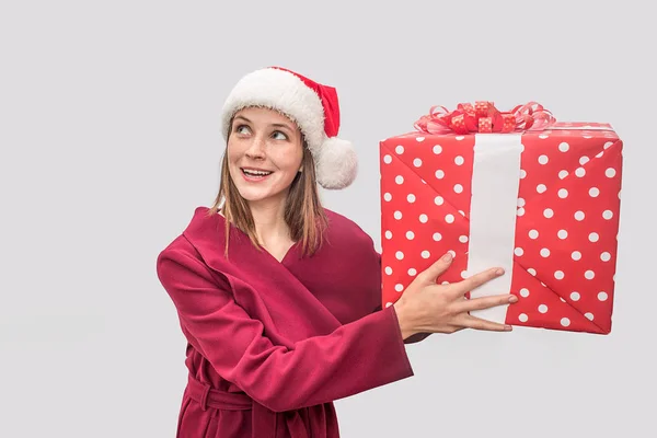 Mujer joven feliz y maravillada se levanta y mira hacia la izquierda. Lleva abrigo rojo y sombrero de Navidad. Modelo sostiene gran regalo en las manos. Ella está emocionada. Aislado sobre fondo gris . — Foto de Stock