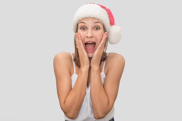 Excited and amazed light-haired woman holds palms on cheeks and screams on camera. She is wondered. Model has Christmas hat on head. Isolated on grey background. — Stock Photo, Image