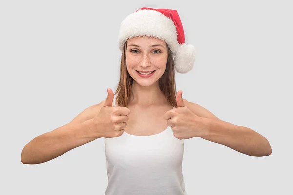 Positive young woman looks on camera and shows her big thumbs up. She wears white t-shirt and red Christmas hat. Model smiles. Isolated on grey background. — Stock Photo, Image