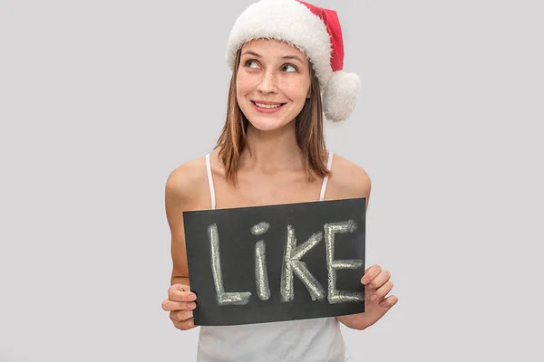 Positive and cheerful young woman looks to side and smiles. She holds plate with written word like. Model wear Christmas hat and white t-shirt. Isolated on grey background. — Stock Photo, Image