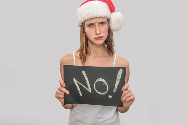 Отрицательная и расстроенная молодая женщина смотрит в камеру. Она несчастна. Модель держит табличку с надписью no Она носит рождественскую шляпу и белую футболку. Изолированный на сером фоне . — стоковое фото