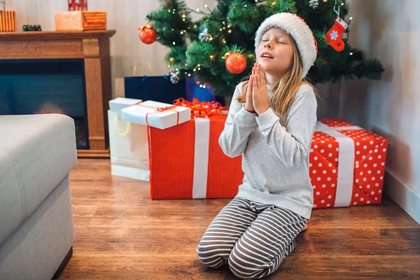 Niña sentada de rodillas y rezando. Mantiene los ojos cerrados y coge las manos. Niño deseando y dando las gracias. Hay cajas con regalos y árbol de Navidad detrás de ella. . — Foto de Stock