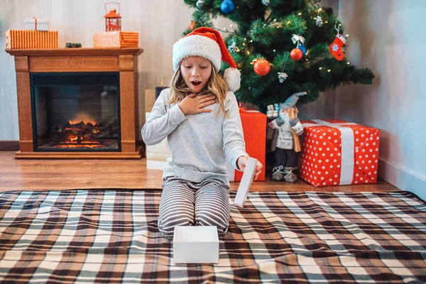 Enfant émerveillé et émerveillé assis sur les genoux et regardant ouvert boîte blanche. Elle tient le dessus en main. Le gamin tient le haut de la boîte à la main. Il y a une cheminée et un arbre de Noël avec des cadeaux derrière elle . — Photo