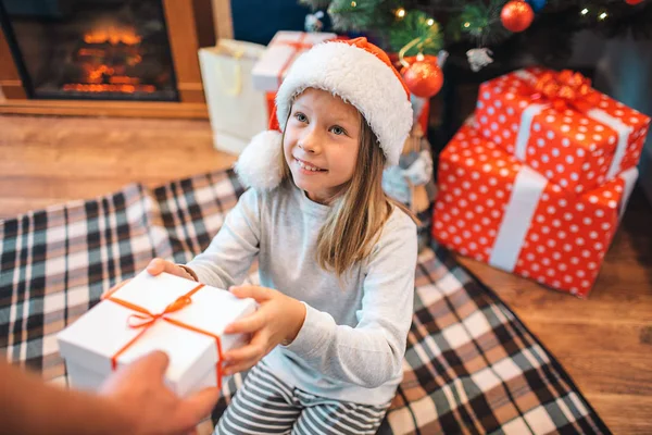 Fille gaie prend boîte avec cadeau et regarde adulte qui lui donne. Elle sourit un peu. La fille est reconnaissante. Elle est assise sur une couverture. Il y a une autre surprise et un sapin de Noël derrière elle . — Photo