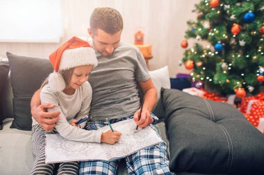 Baba ve kızı birlikte kanepede oturan resim. Onlar çekiyor. İnsanlar ciddi ve konsantre. Genç adam kızını kucaklıyor. Noel ağacı ile dekore edilmiş oda içinde oturuyorlar.